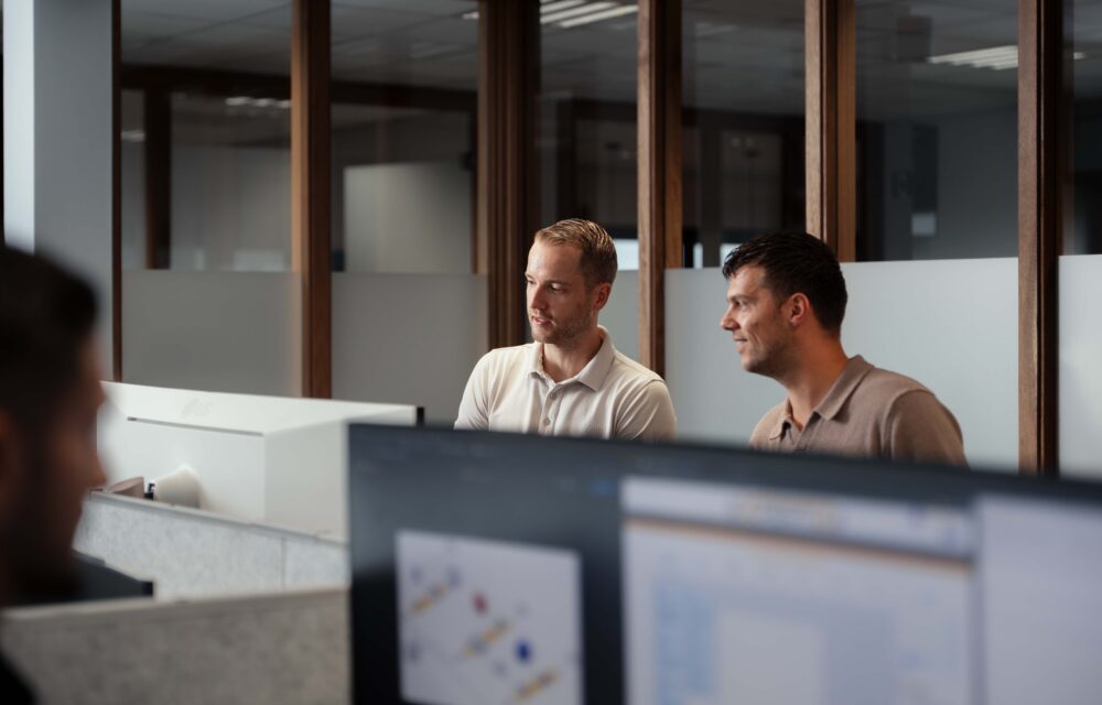 Twee collega's staan naast elkaar achter een bureau en kijken samen naar de computer