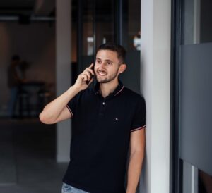Mannelijke collega staat geleund aan een muur en is met zijn telefoon lachend aan het bellen